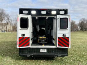An ambulance with its rear doors open reveals an empty stretcher inside. The vehicle is parked on a grassy field, with trees and houses visible in the background.