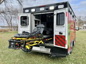 An open ambulance with its back doors wide, showing an empty stretcher extended onto grassy ground. Leafless trees and a cloudy sky are visible in the background.