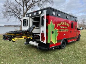 A red and black Maple Bluff Fire Rescue ambulance is parked on grass near a lake, with its back doors open and a yellow stretcher extended outside. Trees and cloudy skies are visible in the background.