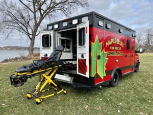 A red Maple Bluff Fire Rescue ambulance with its back doors open is parked on grass near a lake. A yellow and black stretcher is partially extended from the rear of the vehicle. Leafless trees are visible in the background.