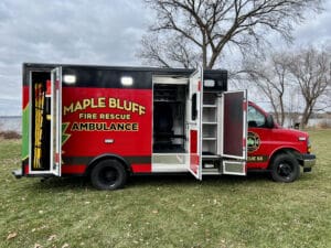 A red Maple Bluff Fire Rescue ambulance with open side doors is parked on grass near leafless trees and a cloudy sky. Emergency equipment is visible inside the vehicle.