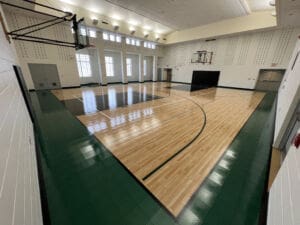 A well-lit indoor basketball court with polished wood flooring, green borders, two basketball hoops, high windows, and white walls. The gym is empty and clean, with overhead lights illuminating the space.