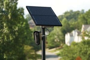 A solar panel mounted on a tall pole with a small security camera attached below it, standing outdoors in a suburban area with trees and houses in the background.