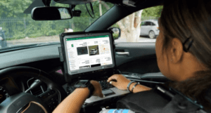 A person in a police uniform sits in a car, using a rugged laptop mounted on the dashboard, searching vehicle information on a screen displaying car images and data.