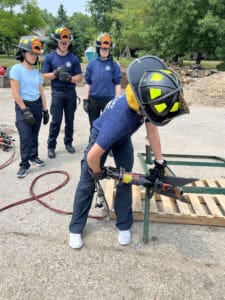 A group of firefighters in uniform and helmets participate in outdoor training. One person uses hydraulic rescue tools on a metal frame while others watch attentively.