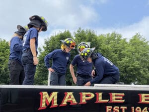 Five firefighters in uniform and helmets stand on top of a fire truck, gathered closely and looking down at something, with green trees and a blue sky in the background. The words MAPLE are visible on the truck.