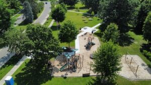 Aerial view of a park with a playground, swings, benches, trees, pathways, and grassy areas on a sunny day.