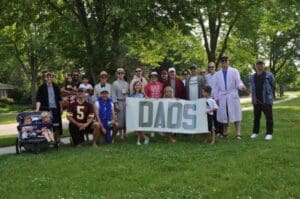 A group of people, including men, women, and children, pose outdoors on grass holding a large sign that says DADS. Trees and a residential street are visible in the background. Some people wear costumes and casual clothes.